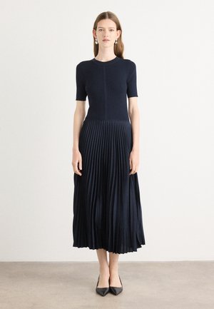 Knit navy dress with short sleeves, featuring a pleated skirt. Smooth top transitions to textured, flowing fabric. Pointed black heels.