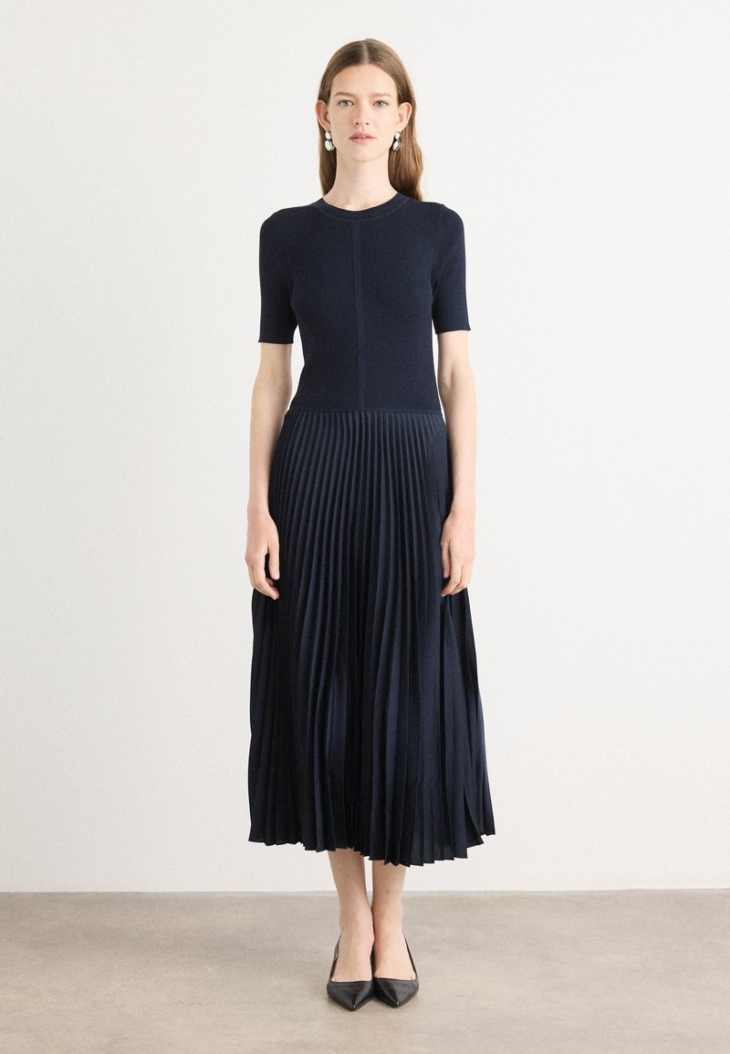 Knit navy dress with short sleeves, featuring a pleated skirt. Smooth top transitions to textured, flowing fabric. Pointed black heels.
