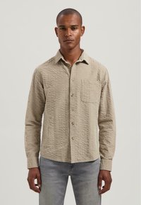 Hombre vestido con una camiseta de botones de manga larga beige texturizada con un bolsillo en el pecho y jeans gris claro, de pie frente a un fondo liso.