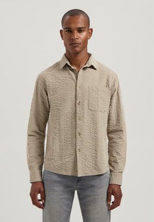 Hombre vestido con una camiseta de botones de manga larga beige texturizada con un bolsillo en el pecho y jeans gris claro, de pie frente a un fondo liso.