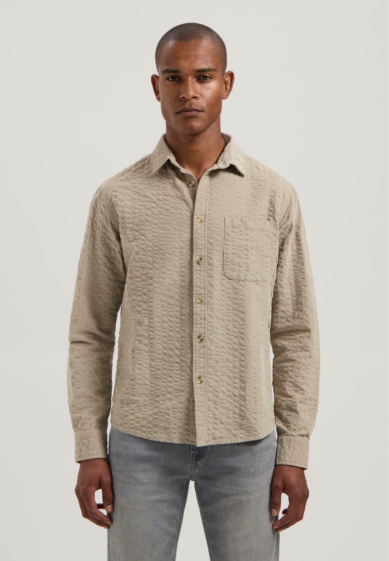 Hombre vestido con una camiseta de botones de manga larga beige texturizada con un bolsillo en el pecho y jeans gris claro, de pie frente a un fondo liso.