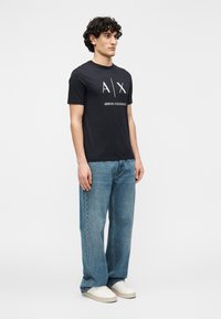 Μαύρο μπλουζάκι με κοντά μανίκια και λευκό λογότυπο «A|X ARMANI EXCHANGE», συνδυασμένο με ανοιχτό μπλε τζιν παντελόνι φαρδιάς γραμμής και λευκά αθλητικά παπούτσια. Μινιμαλιστικός σχεδιασμός.