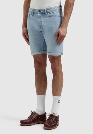 Man draagt lichtblauwe denim shorts, witte sportsokken en bruine leren bootschoenen, staat met handen ontspannen langs zijn zij.