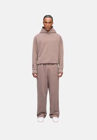 Sudadera color marrón claro y pantalones a juego, hechos de una tela suave, con un corte holgado, puños acanalados y un detalle de logo en los pantalones. Zapatillas blancas.