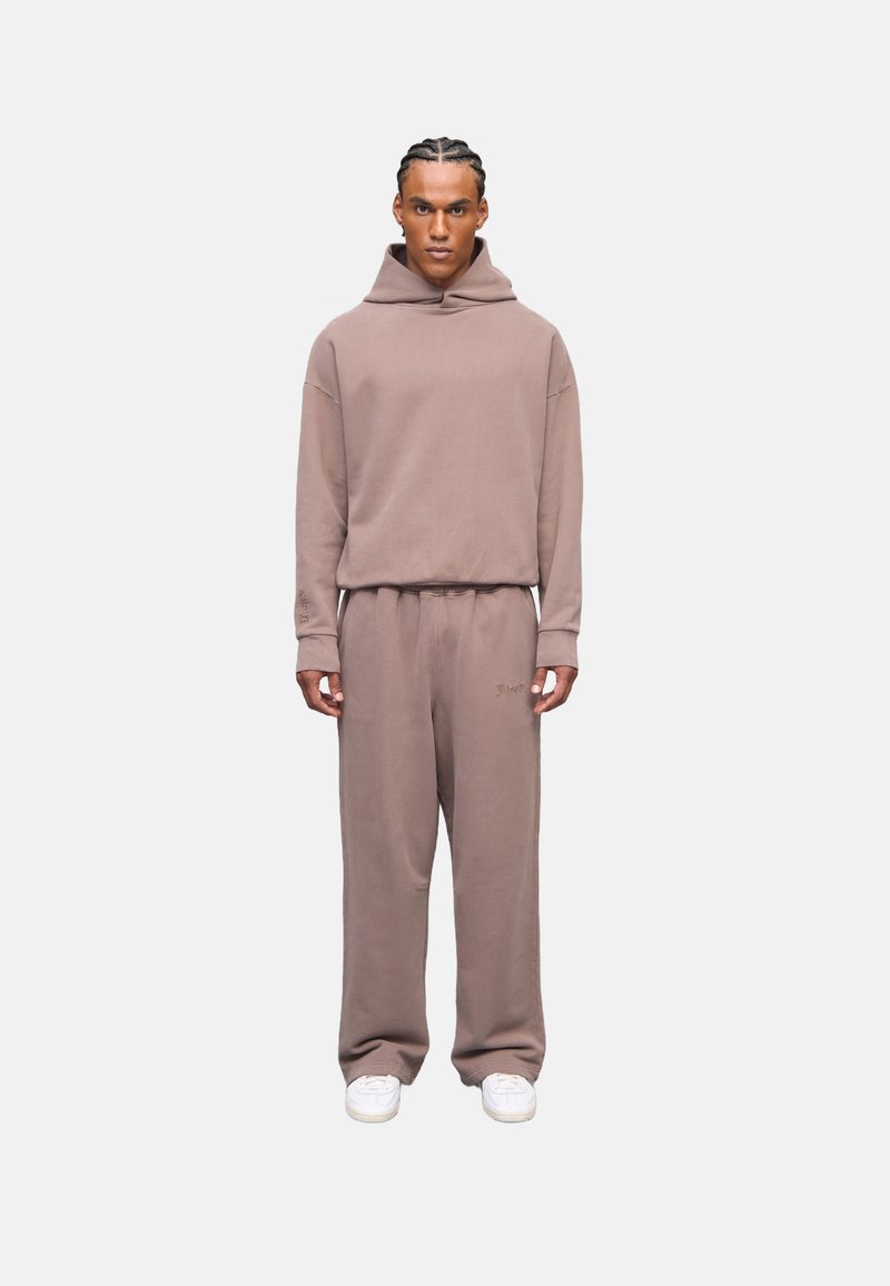 Sudadera color marrón claro y pantalones a juego, hechos de una tela suave, con un corte holgado, puños acanalados y un detalle de logo en los pantalones. Zapatillas blancas.