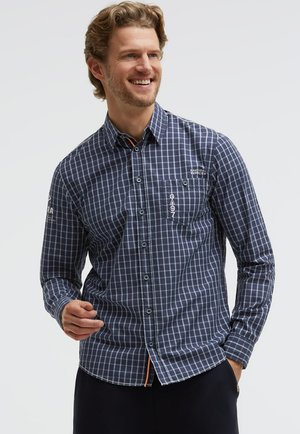 Smiling man wearing navy blue checkered button-up shirt with pocket and white nautical text, standing against white background.