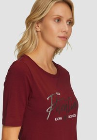 Bordeaux katoenen t-shirt met korte mouwen en een ronde hals, met een zilveren tekstafbeelding die "JOUW potentieel KENT geen BEPERKINGEN" zegt.