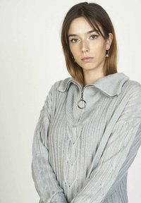 Joven mujer con cabello castaño hasta los hombros que lleva un suéter de punto en color gris claro con cremallera y anilla, mirando a la cámara.