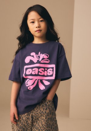 Jeune fille aux longs cheveux foncés portant un t-shirt violet foncé avec l'inscription rose « oasis » et un pantalon imprimé léopard, une main dans la poche.