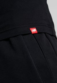 Tejido atlético negro con una textura suave, que presenta una prominente etiqueta con el logo rojo de New Balance en la cinturilla. Diseño de ajuste relajado con pliegues.