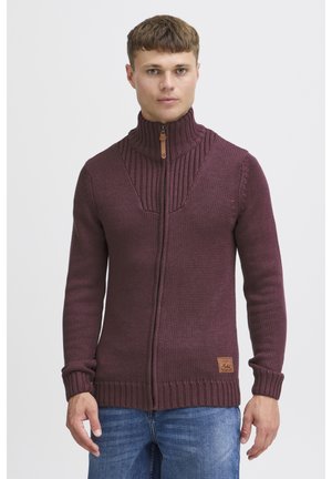 Maglione lavorato a maglia color bordeaux con colletto alto e chiusura con zip. Presenta una texture a coste sulle spalle e sui polsini, con una toppa del logo vicino all'orlo.