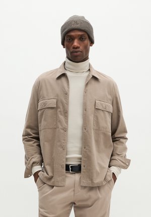 Mann trägt ein beiges Hemd mit Knöpfen über einem weißen Rollkragenpullover, beige Hose, schwarzen Gürtel und graue Strickmütze mit gestickten Buchstaben.
