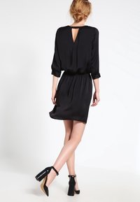Vestido negro con un corte holgado, mangas largas y un escote en forma de lágrima en la espalda. Textura suave y cintura ajustada. Combinado con zapatos de tacón alto negros.