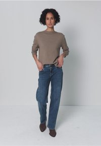Bruine gebreide trui met een boothals, gecombineerd met relaxed blauwe jeans en bruine pumps met een spitse neus. Model staat met handen in de zij.