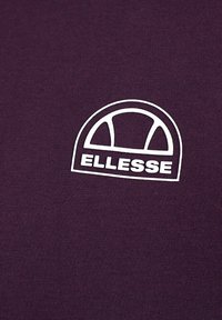 Camiseta de algodón morada con un logo blanco de forma redondeada y el texto "ELLESSE" debajo, sobre un fondo de tejido texturizado.