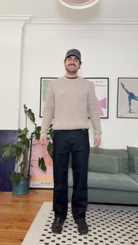 Camisola bege clara canelada, jeans escuros e sapatos pretos. Ambiente interior com planta verde e arte emoldurada em paredes brancas.