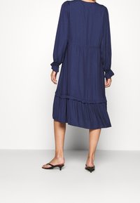 Tmavě modré šaty s dlouhým rukávem, vertikálními sklady, řaseným lemem a elastickými manžetami. Lehká tkanina volně splývá a nabízí uvolněný střih.