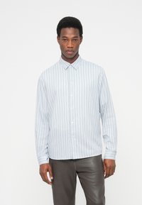 Άνδρας με κοντά σγουρά μαλλιά, φοράει λευκό πουκάμισο με μακριά μανίκια και λεπτές κάθετες ρίγες, καθώς και σκούρο παντελόνι, στέκεται μπροστά σε μια απλή φόντο.