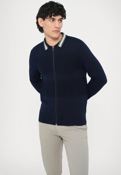 Námornícky modrý sveter na zips s pruhovaným golierom. Má textúrované telo a priliehavý strih. Skombinujte s svetlo béžovými nohavicami pre kontrast.