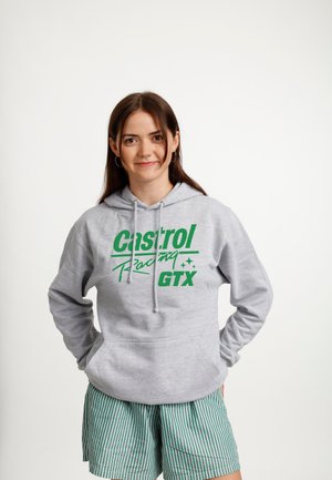 Junge Frau mit langen braunen Haaren trägt einen grauen Kapuzenpullover mit grünem "Castrol Racing GTX"-Logo und grün gestreifte Shorts, steht mit den Händen in den Taschen.