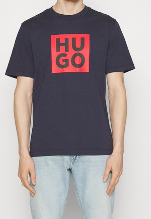 Marineblaues Baumwoll-T-Shirt mit einer großen roten quadratischen Grafik und dem Text "HUGO" in schwarzen Buchstaben auf der Vorderseite. Kurze Ärmel und Rundhalsausschnitt.