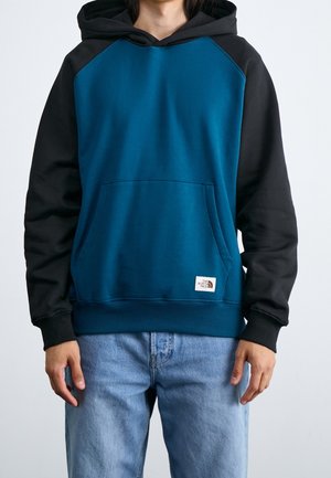 Persona che indossa una felpa con cappuccio blu e nera, con tasca frontale, e jeans blu chiaro, in piedi contro uno sfondo neutro.