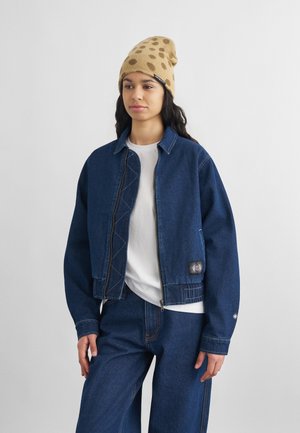 Jauna moteris dėvi gelsvai taškuotą megztą kepuraitę, baltą marškinėlį, tamsiai mėlyną džinsinę striukę su lopu ir atitinkančias džinsines kelnes, stovinti priešais paprastą fono sieną.