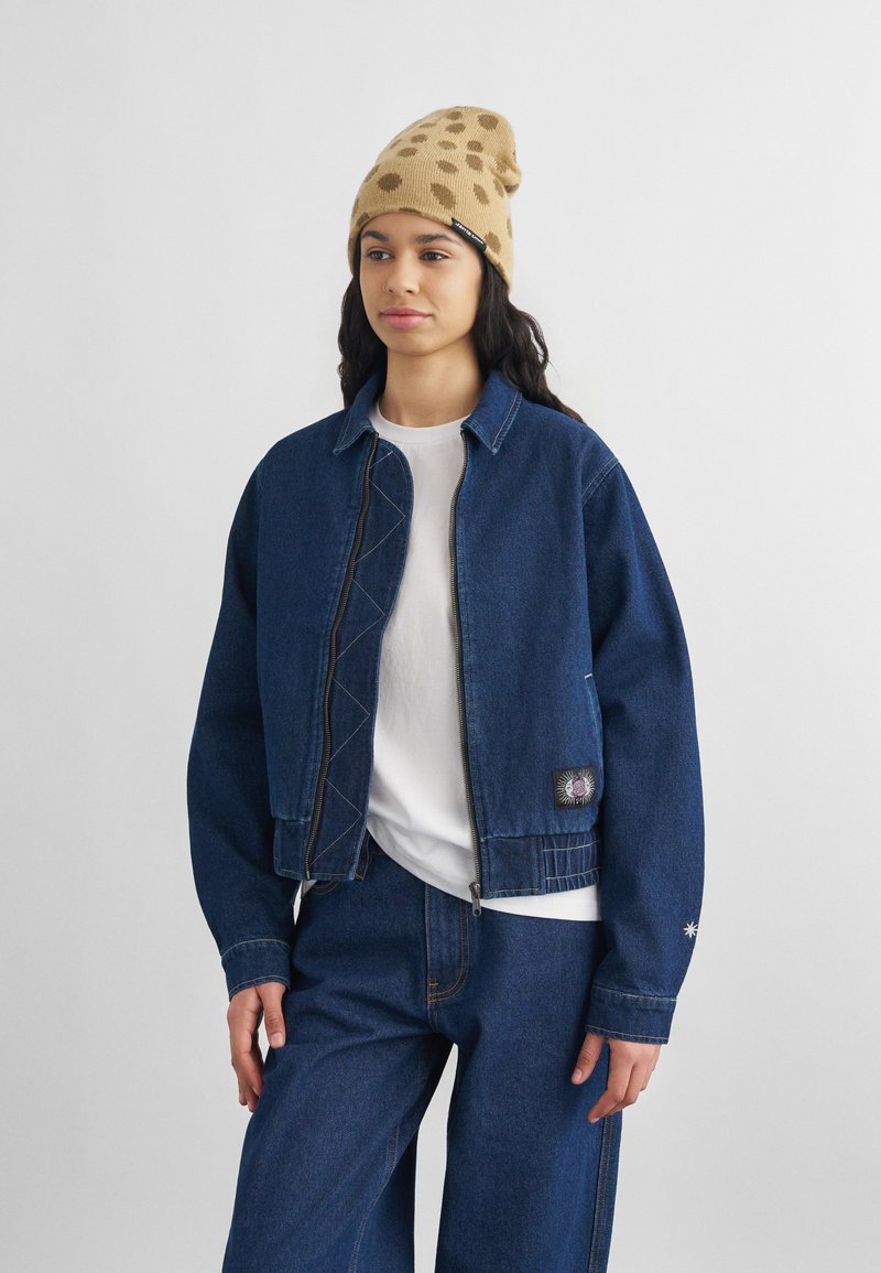 Νεαρή γυναίκα που φοράει καφέ σκούφο με σχέδια, λευκό t-shirt, σκούρο μπλε τζιν μπουφάν με πάτσ, και τζιν παντελόνι που ταιριάζει, στέκεται μπροστά σε ένα απλό φόντο.