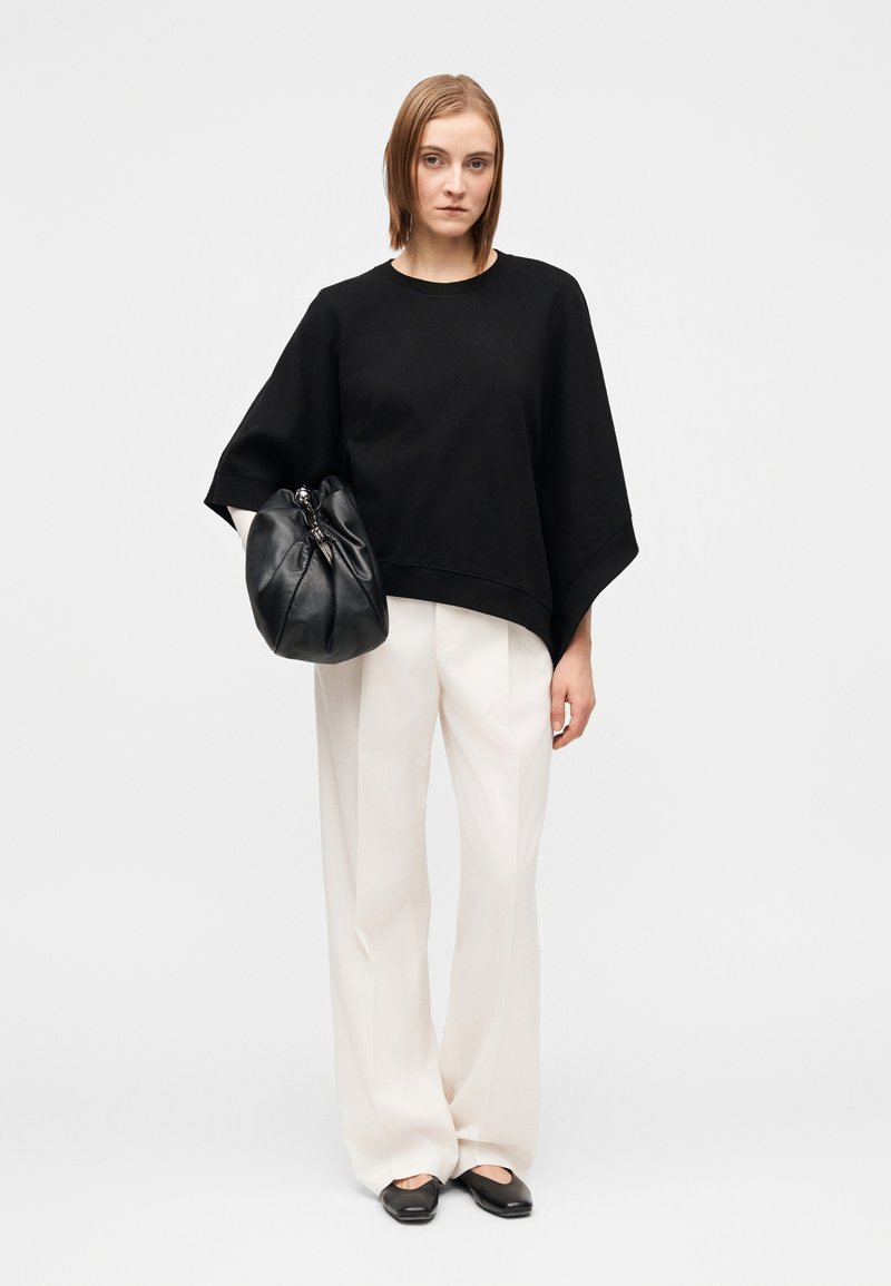 Sweatshirt noir surdimensionné avec manches larges, associé à un pantalon large crème. Accessoires comprenant un sac à main froncé noir et des chaussures plates.