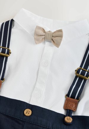 Camicia bianca con tessuto strutturato, papillon beige e bretelle blu navy con strisce bianche, dettagli in pelle marrone e bottoni.