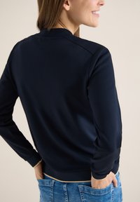 Femme portant un pull à manches longues bleu marine avec des bordures beiges, les mains dans les poches, souriante, vue de dos et de profil sur un fond neutre.
