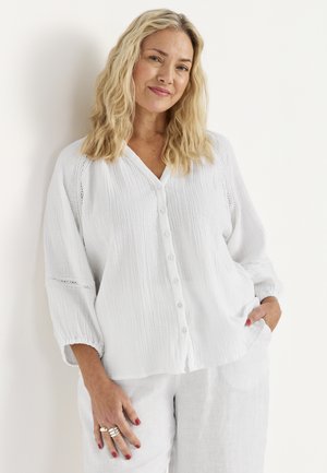 Blondynka o falowanych włosach, ubrana w białą koszulę na guziki i białe spodnie, stojąca z jedną ręką w kieszeni na białym tle.