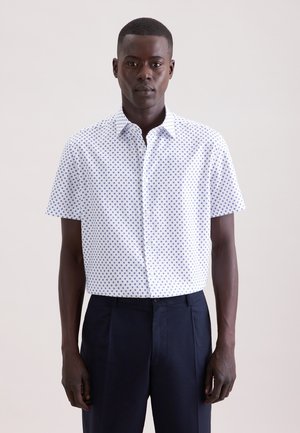 Mann trägt ein kurzärmeliges weißes Hemd mit blauen Punkten und dunkelblaue Hose, steht vor einem einfarbigen hellen Hintergrund.