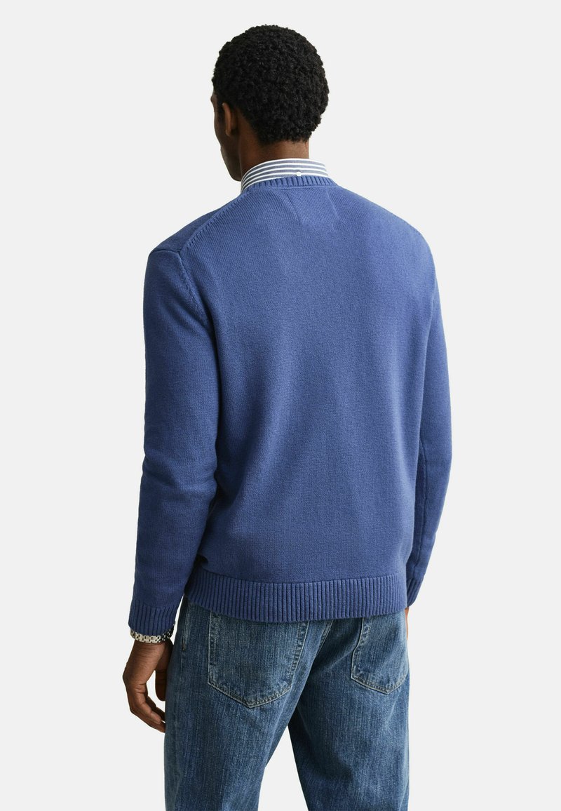 Mann trägt blauen Strickpullover über gestreiftem Hemd mit Kragen und blauen Jeans, von hinten vor einfarbigem Hintergrund gesehen.