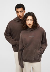 Δύο μοντέλα φορούν υπερμεγέθη σκουρόχρωμα καφέ φούτερ με κουκούλα, κατασκευασμένα από απαλό, υφασμάτινο υλικό με υφή, που διαθέτουν μπροστινές τσέπες και το κείμενο «URTRN» σε λευκή κεντητική.