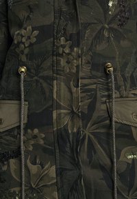 Bunda s maskáčovým vzorem ve tmavě zelené látce s květinovými motivy, dekorativními flitry, nastavitelnými šňůrkami a dvěma předními kapsami.