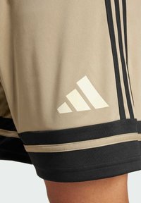 Béžové sportovní šortky s černými detaily, které mají tři černé pruhy po straně a bílé logo Adidas na dolní části levé nohy.