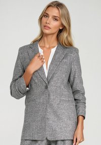 Szary żakiet z tweedu z pojedynczym zapięciem na guzik, klapami, oraz dwoma przednimi kieszeniami; charakteryzuje się teksturowanym, tkanym materiałem.