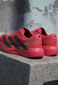 Ζευγάρι κόκκινα παπούτσια Adidas για τρέξιμο με μαύρες ρίγες τοποθετημένα σε γκρι πέτρινη επιφάνεια με φόντο υφή τοίχου.