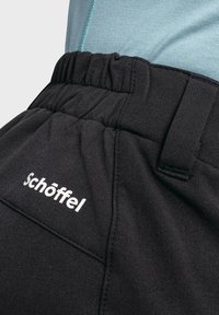 Schwarze Hose mit elastischem Bund, ausgestattet mit einem weißen Logo, glatter Textur und taillierter Passform. Teil eines geschichteten Outfits mit einem blauen Oberteil.