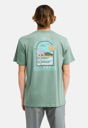Persona che indossa una t-shirt verde con una scena pastello di spiaggia, palme, sole e il testo "Billabong Adventure Division" sulla schiena.