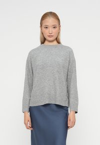 Pulover gri din tricot cu model rib, cu decolteu rotund, mâneci lungi și accente cu nasturi pe laterale, purtat peste o fustă netedă de culoare albastru marin.
