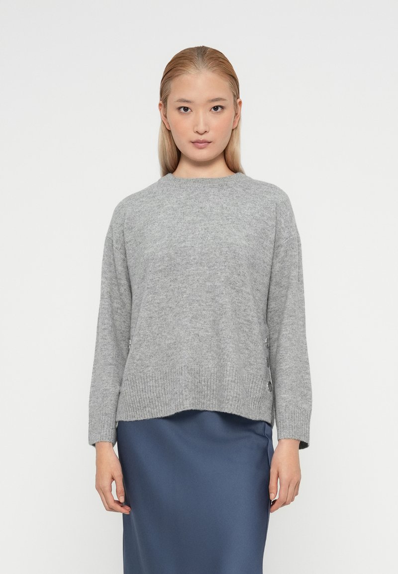 Pulover gri din tricot cu model rib, cu decolteu rotund, mâneci lungi și accente cu nasturi pe laterale, purtat peste o fustă netedă de culoare albastru marin.