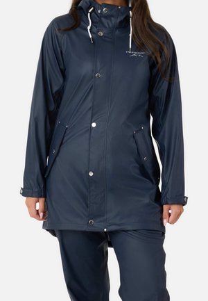 Femme portant un imperméable bleu marine avec capuche, boutons-pression et poches avant, assorti à un pantalon bleu marine.