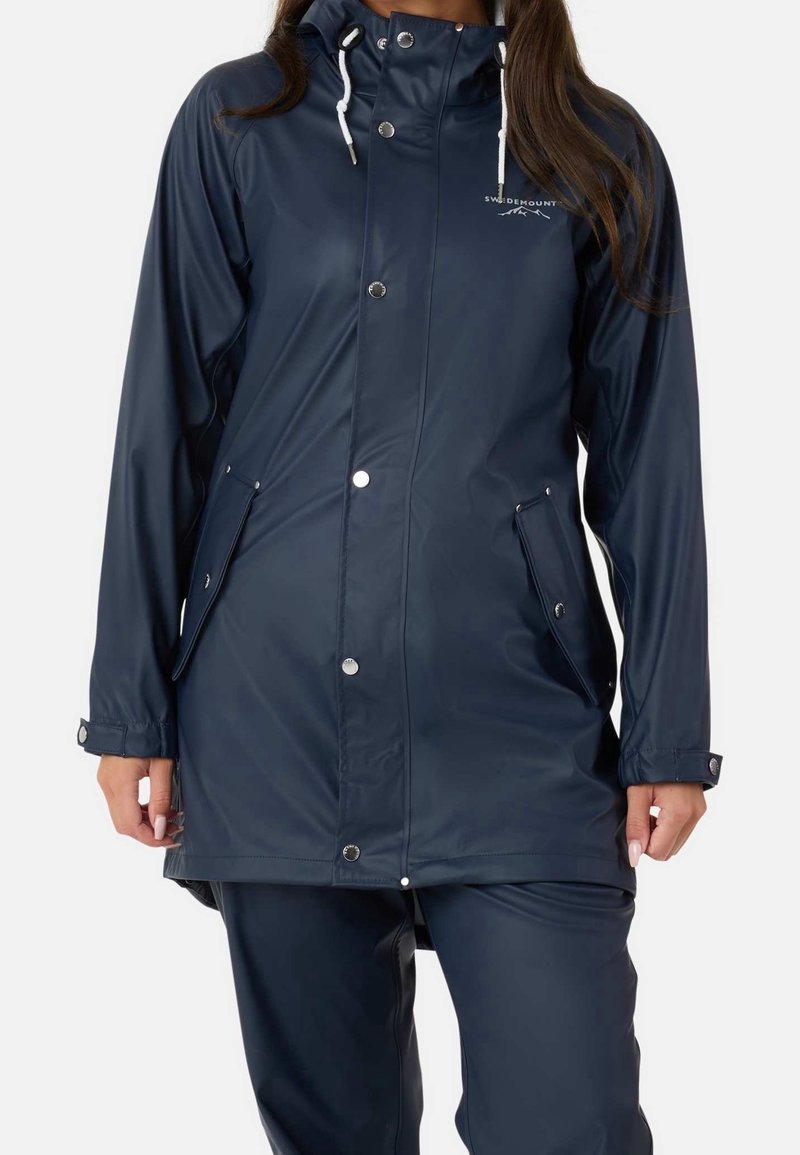 Femme portant un imperméable bleu marine avec capuche, boutons-pression et poches avant, assorti à un pantalon bleu marine.