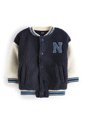 Veste varsity sherpa bleu marine et crème avec boutons pression, poignets et col rayés, et un écusson lettre "N" bleu sur la poitrine.
