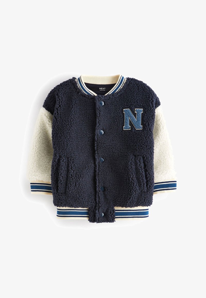 Veste varsity sherpa bleu marine et crème avec boutons pression, poignets et col rayés, et un écusson lettre "N" bleu sur la poitrine.