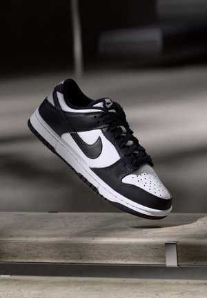 Baskets Nike Dunk noir et blanc avec lacets, semelle intermédiaire et logo noirs, positionnées au-dessus d'une surface en béton avec un arrière-plan flou.