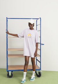 Camiseta de gran tamaño en color lila con un gráfico de arcoíris, combinada con una gorra blanca, calcetines blancos y zapatillas de color teal, al lado de una estantería metálica azul.