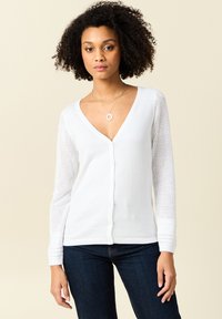 Vit cardigan med V-urringning och långa ärmar som har ett stickat mönster. Den har knappar framtill och ribbad fålldetalj.